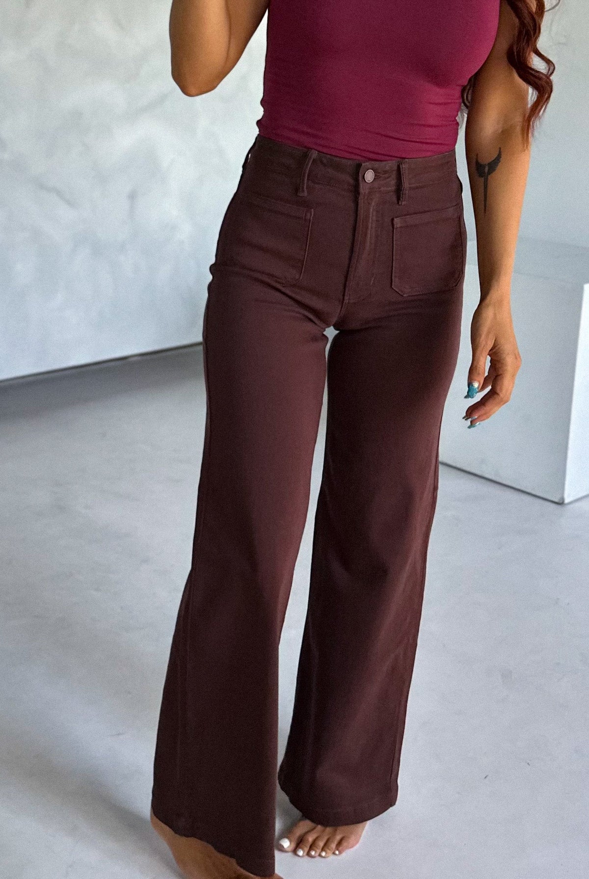 Woman taking a mirror selfie wearing a maroon tank top and brown pants in a minimalistic room.