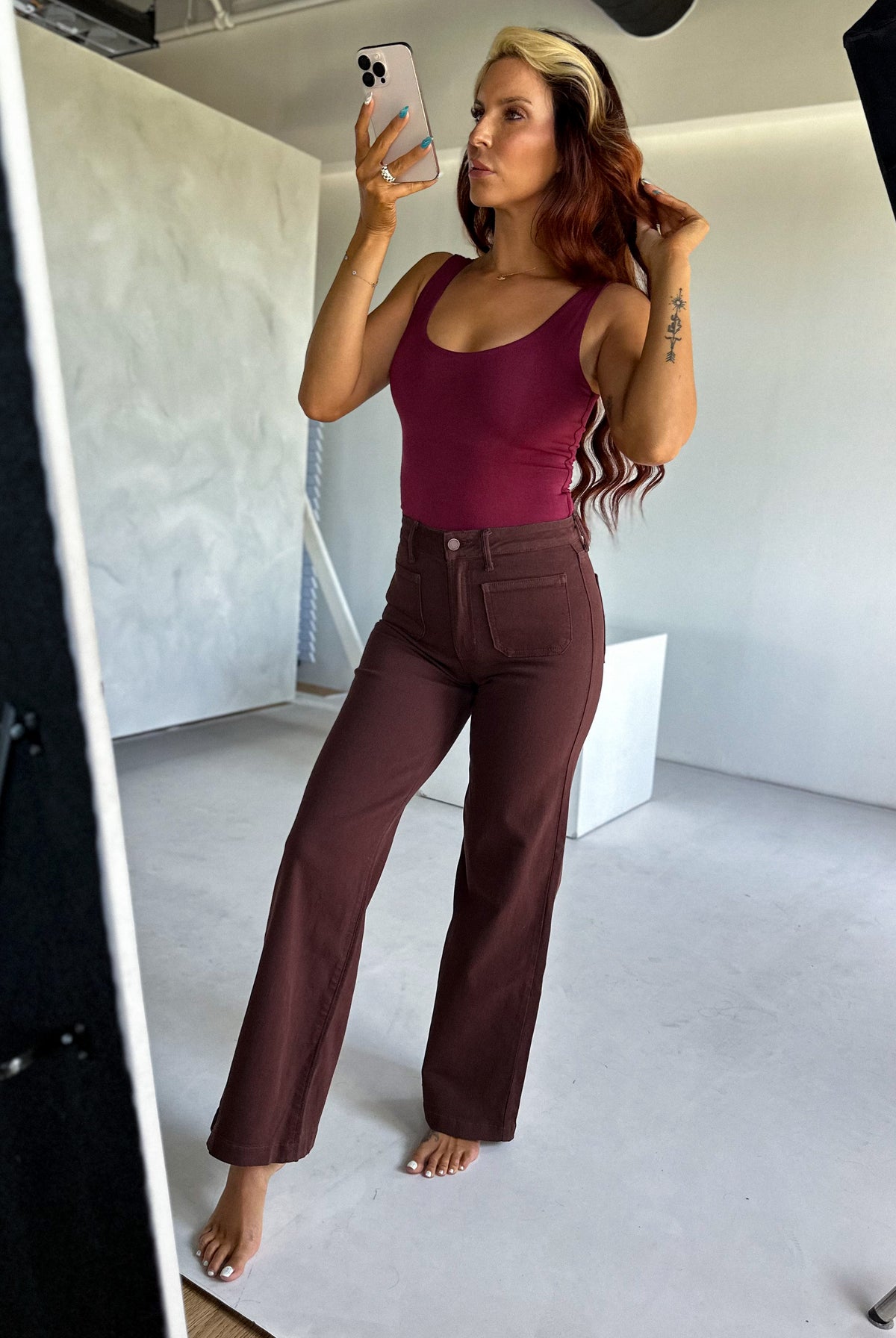 Woman taking a mirror selfie wearing a burgundy top and brown pants in a studio setting.
