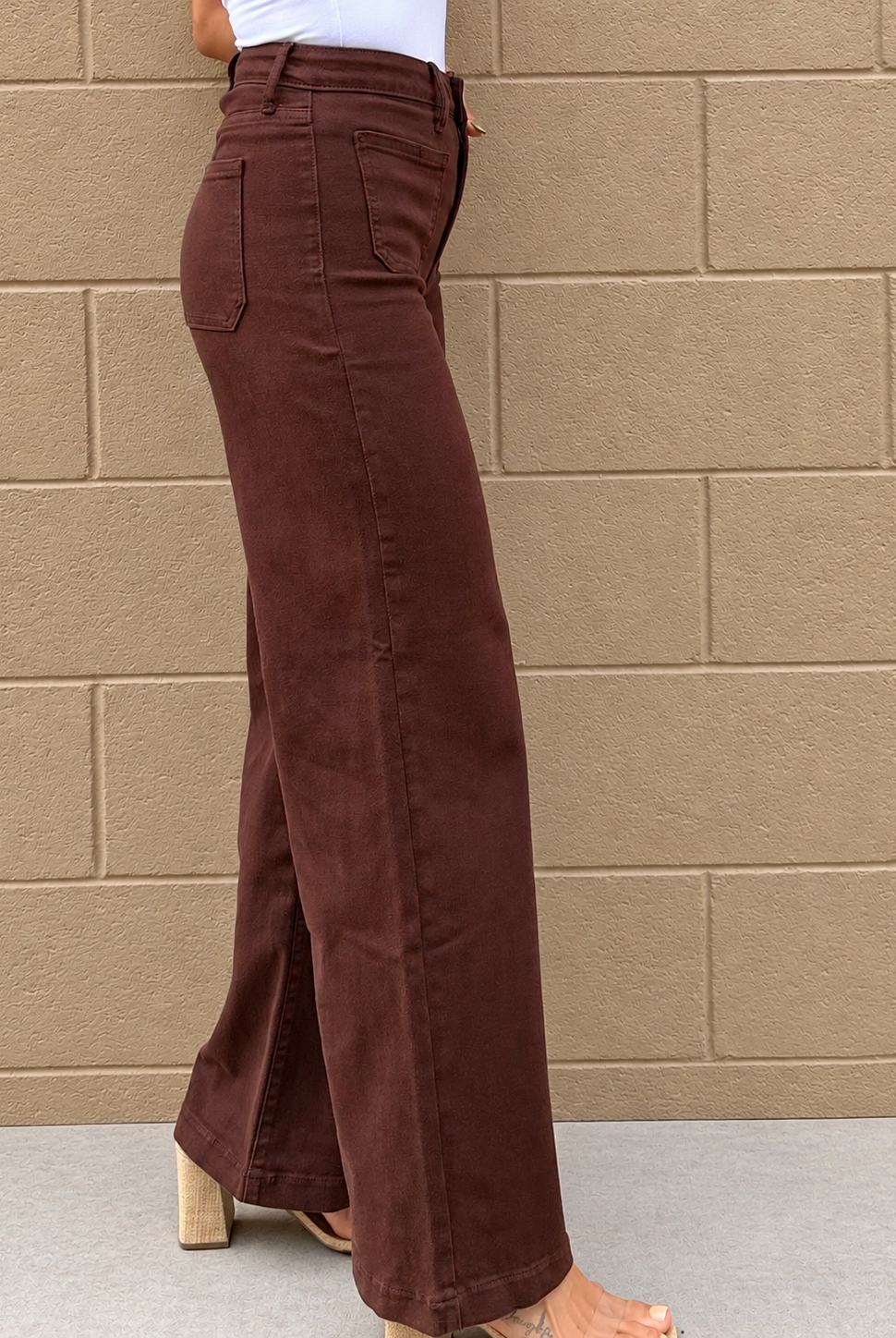 A person wearing high-waisted, wide-leg brown pants with a side slit, paired with a white top and block-heel sandals, standing against a tan brick wall.