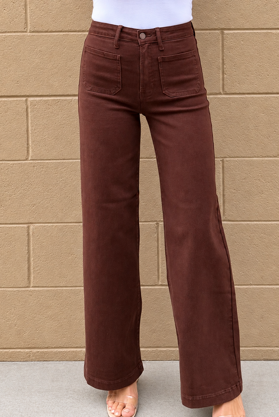 A person wearing dark brown wide-leg jeans with a white top, standing against a tan brick wall.