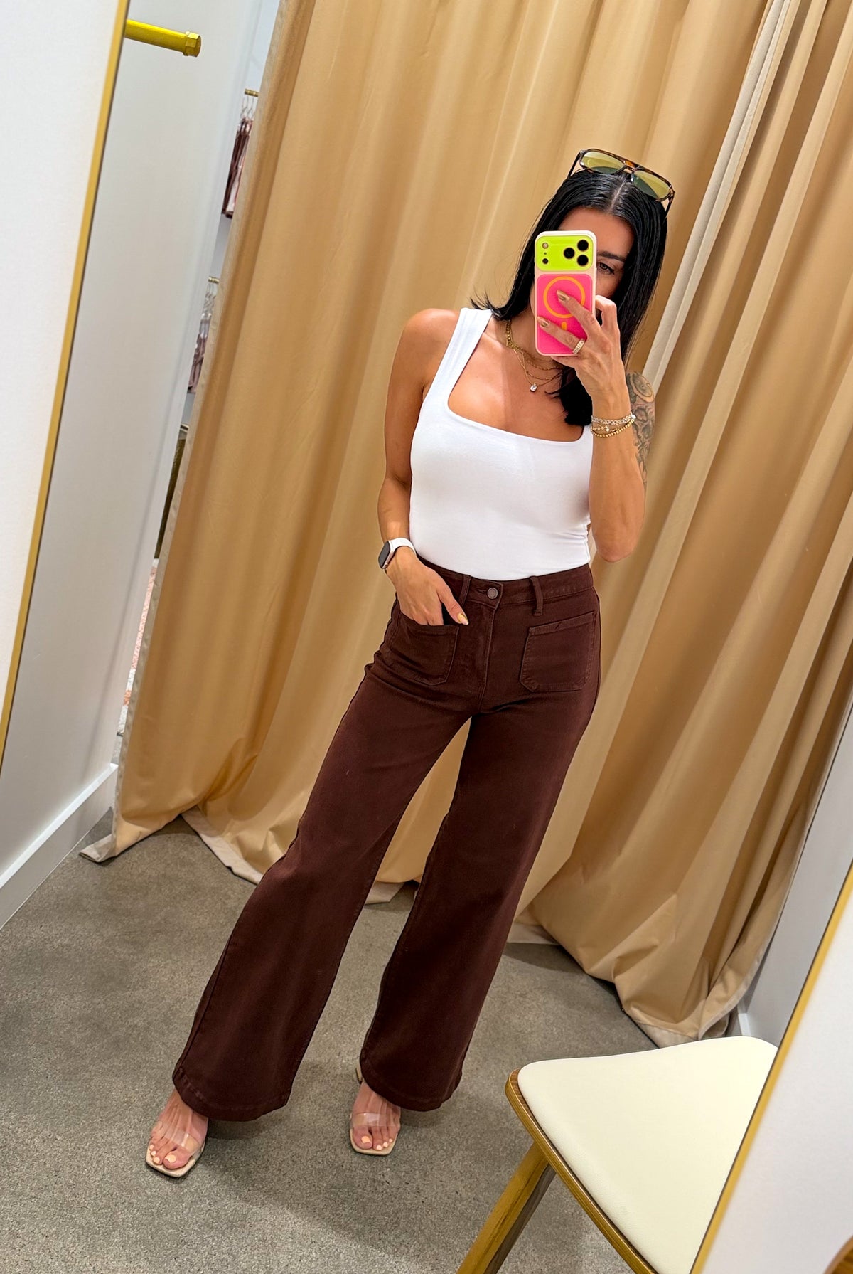 A woman takes a mirror selfie in a fitting room wearing a white tank top and dark brown wide-leg pants. She is holding a pink phone, with beige curtains and a gray carpet in the background.
