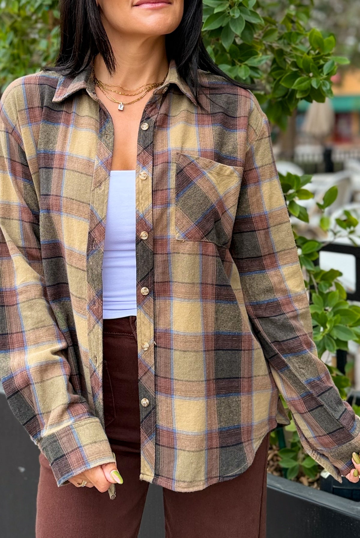 A woman wearing a long-sleeve button-up plaid flannel shirt in beige, brown, and muted blue, layered over a white tank top and brown pants, photographed outdoors with green foliage in the background.