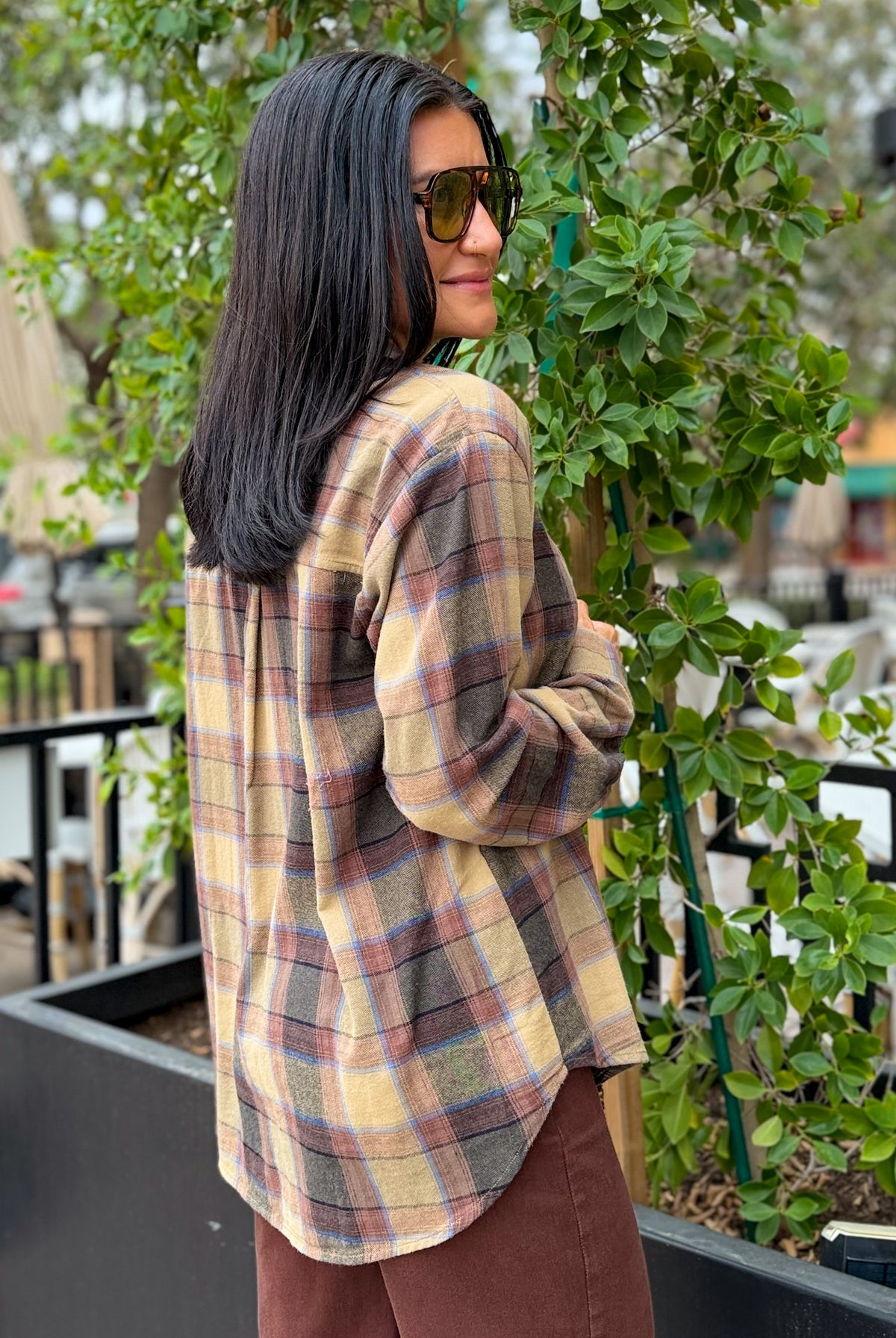 A woman with long black hair and sunglasses, wearing a beige and brown plaid flannel shirt, standing outdoors next to a green plant.