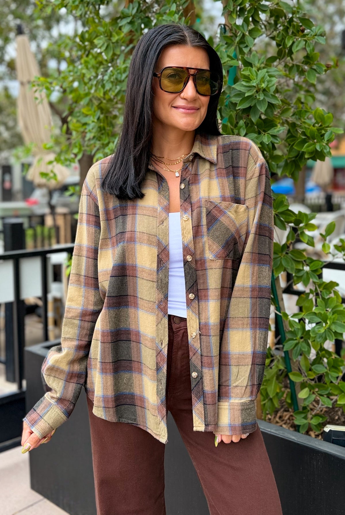 A woman wearing an oversized long-sleeve plaid button-up shirt in beige, brown, and muted red tones, styled over a white top and brown pants, standing outdoors in front of greenery.