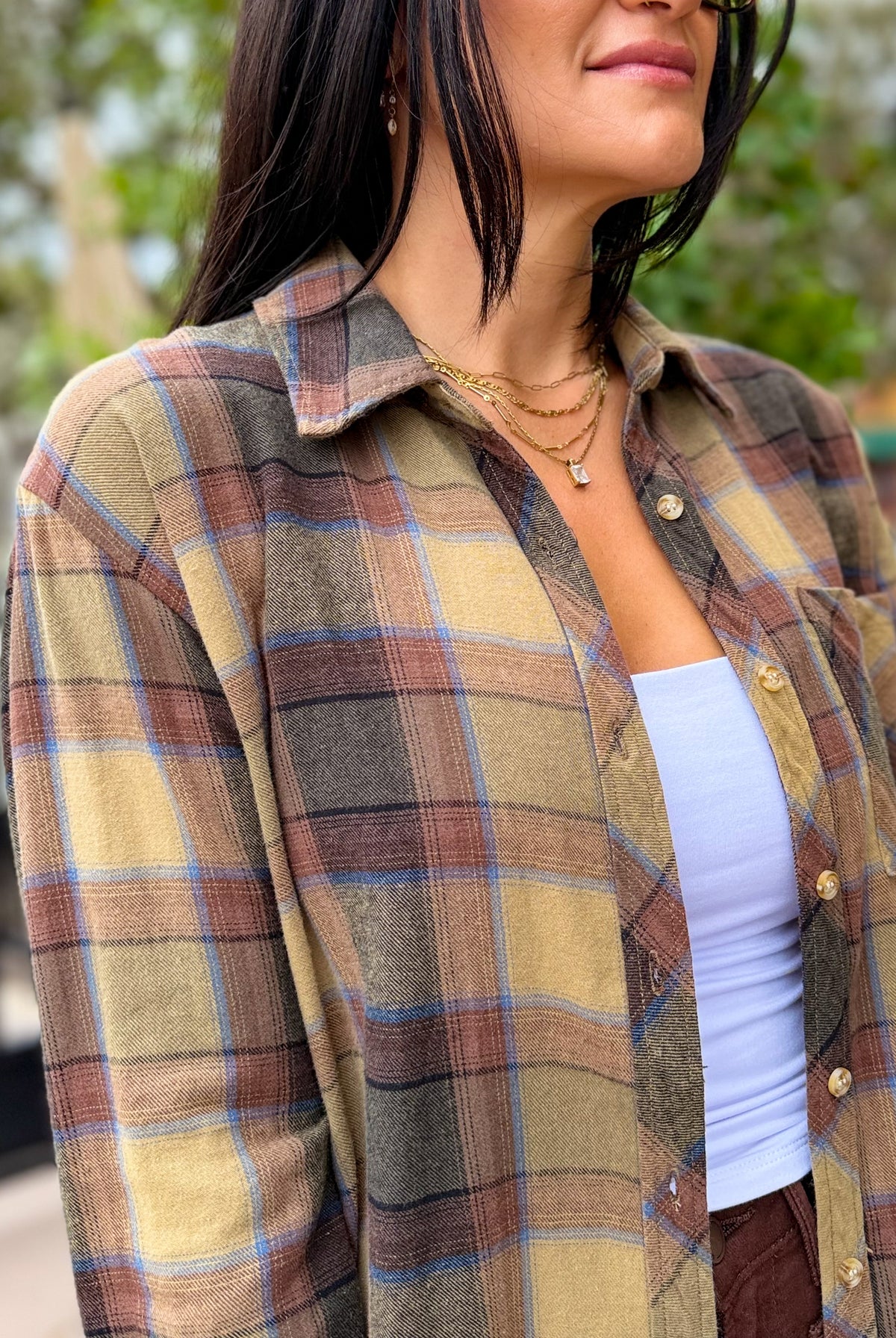 A woman wearing a long-sleeve plaid button-up shirt in muted earthy tones of beige, brown, gray, and light blue, layered over a white tank top.