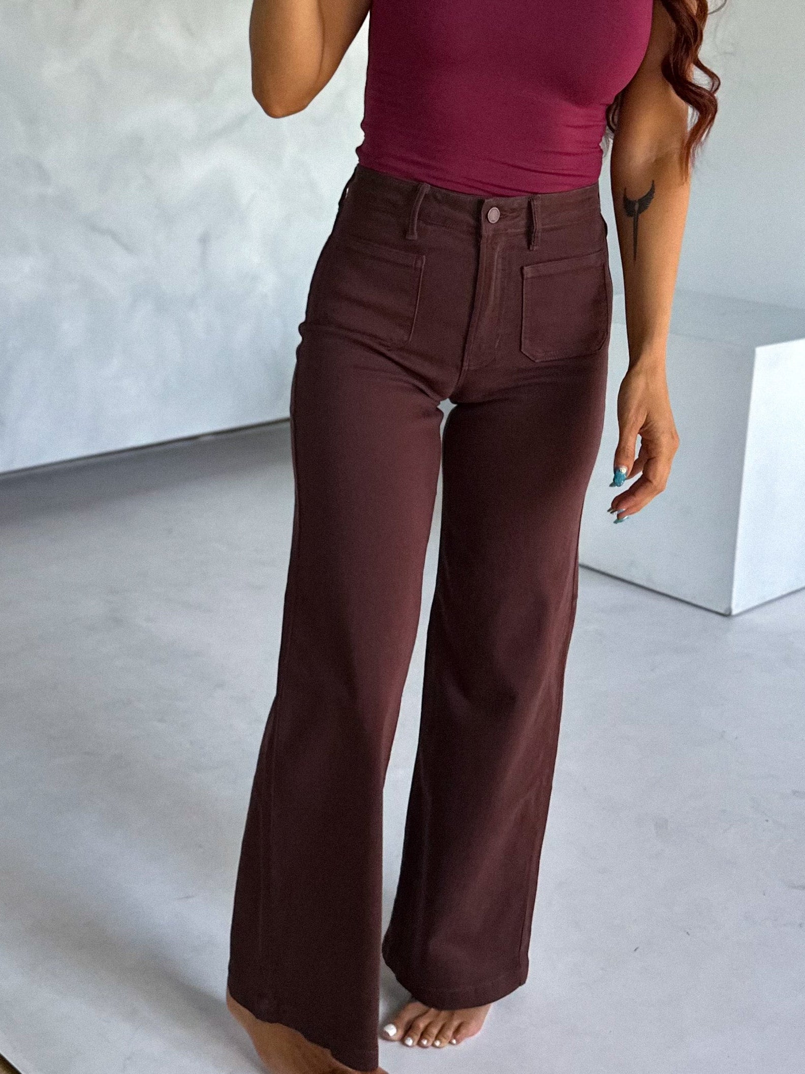 Woman taking a mirror selfie wearing a maroon tank top and brown pants in a minimalistic room.