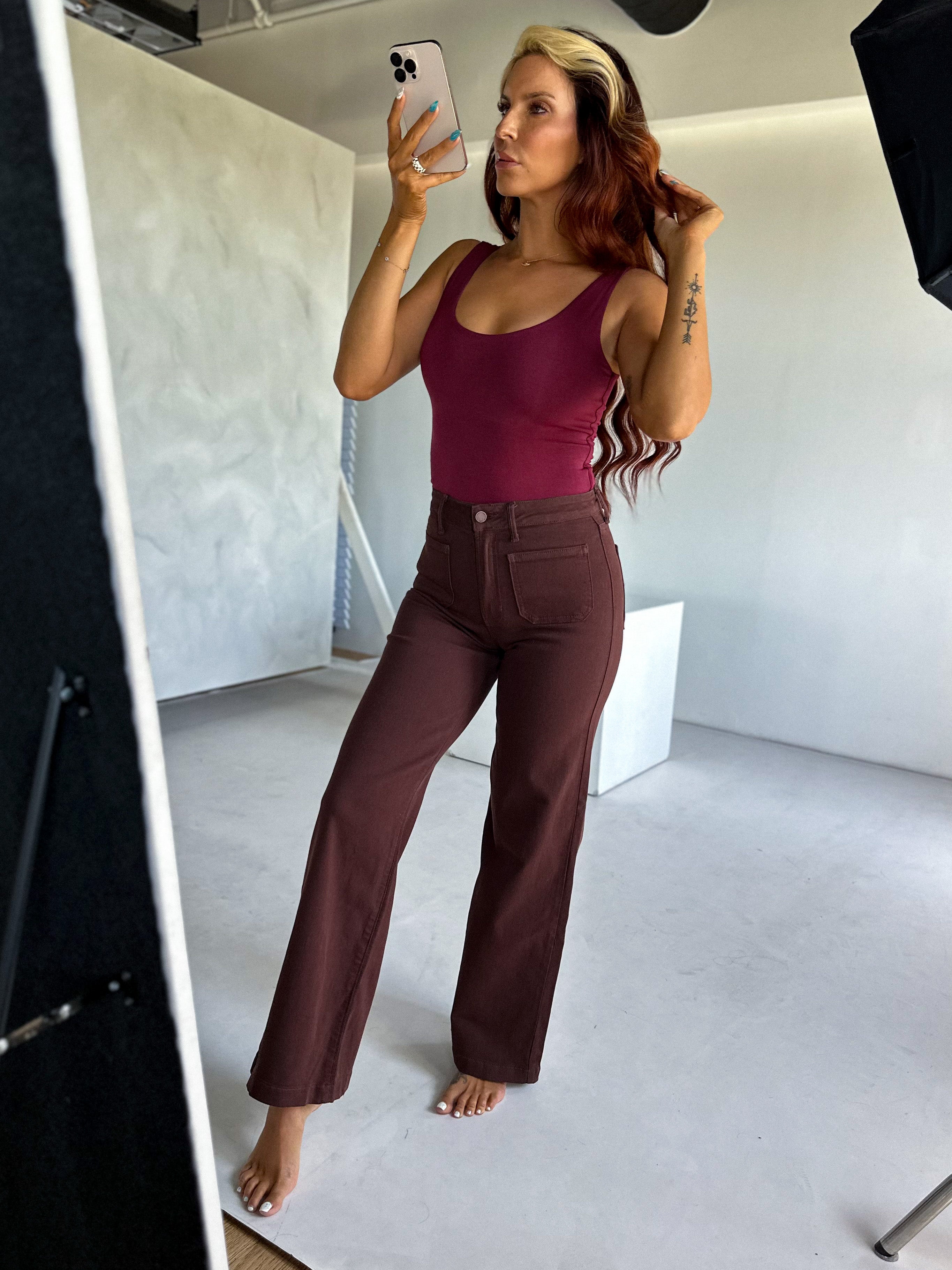 Woman taking a mirror selfie wearing a burgundy top and brown pants in a studio setting.