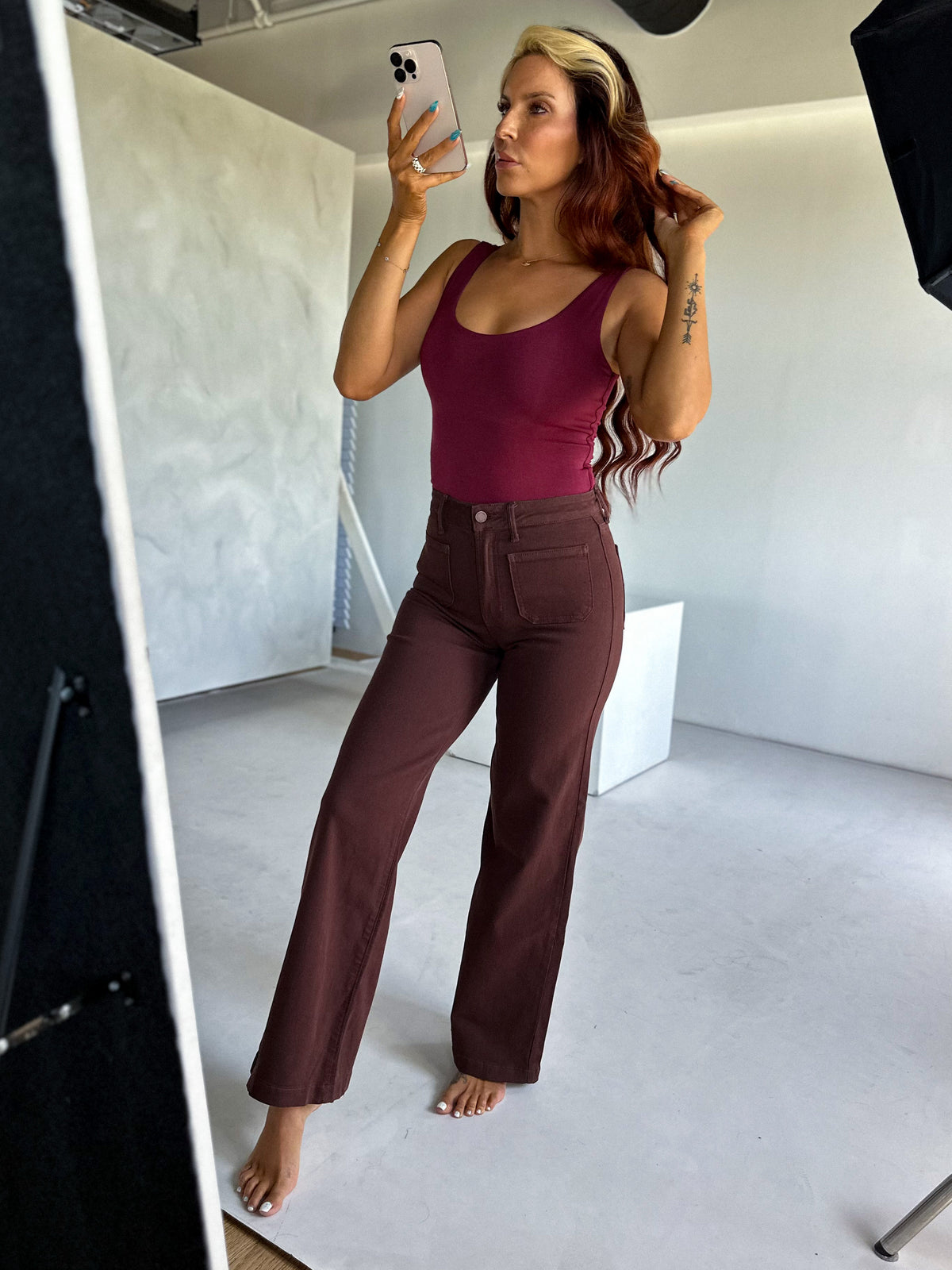 Woman taking a mirror selfie wearing a burgundy top and brown pants in a studio setting.