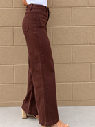 A person wearing high-waisted, wide-leg brown pants with a side slit, paired with a white top and block-heel sandals, standing against a tan brick wall.