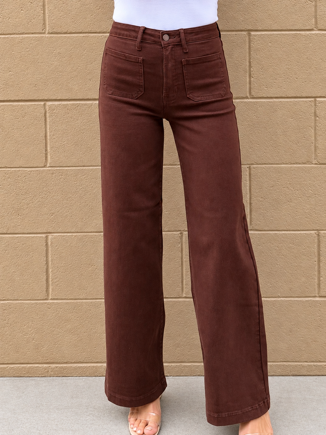 A person wearing dark brown wide-leg jeans with a white top, standing against a tan brick wall.