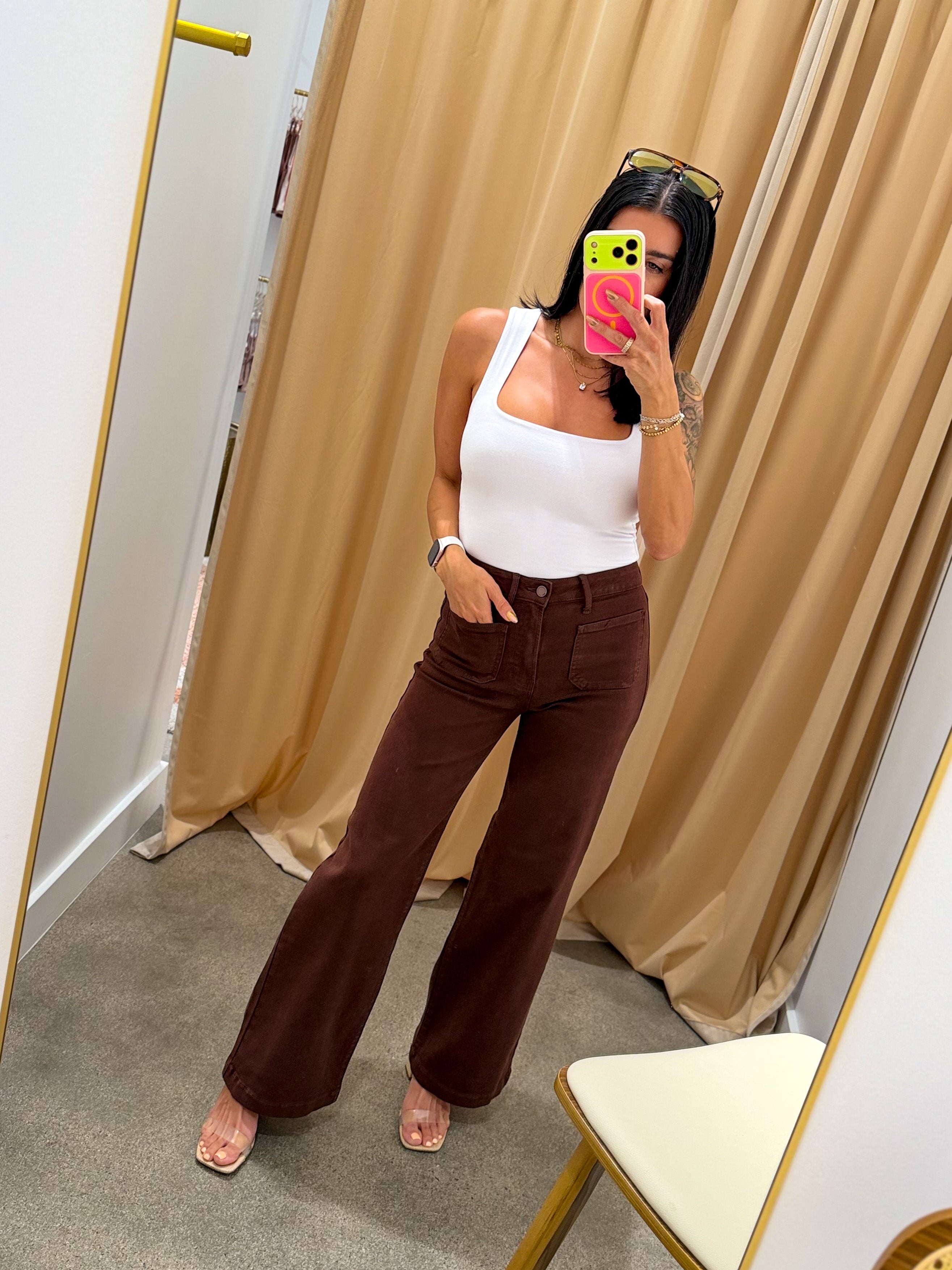 A woman takes a mirror selfie in a fitting room wearing a white tank top and dark brown wide-leg pants. She is holding a pink phone, with beige curtains and a gray carpet in the background.