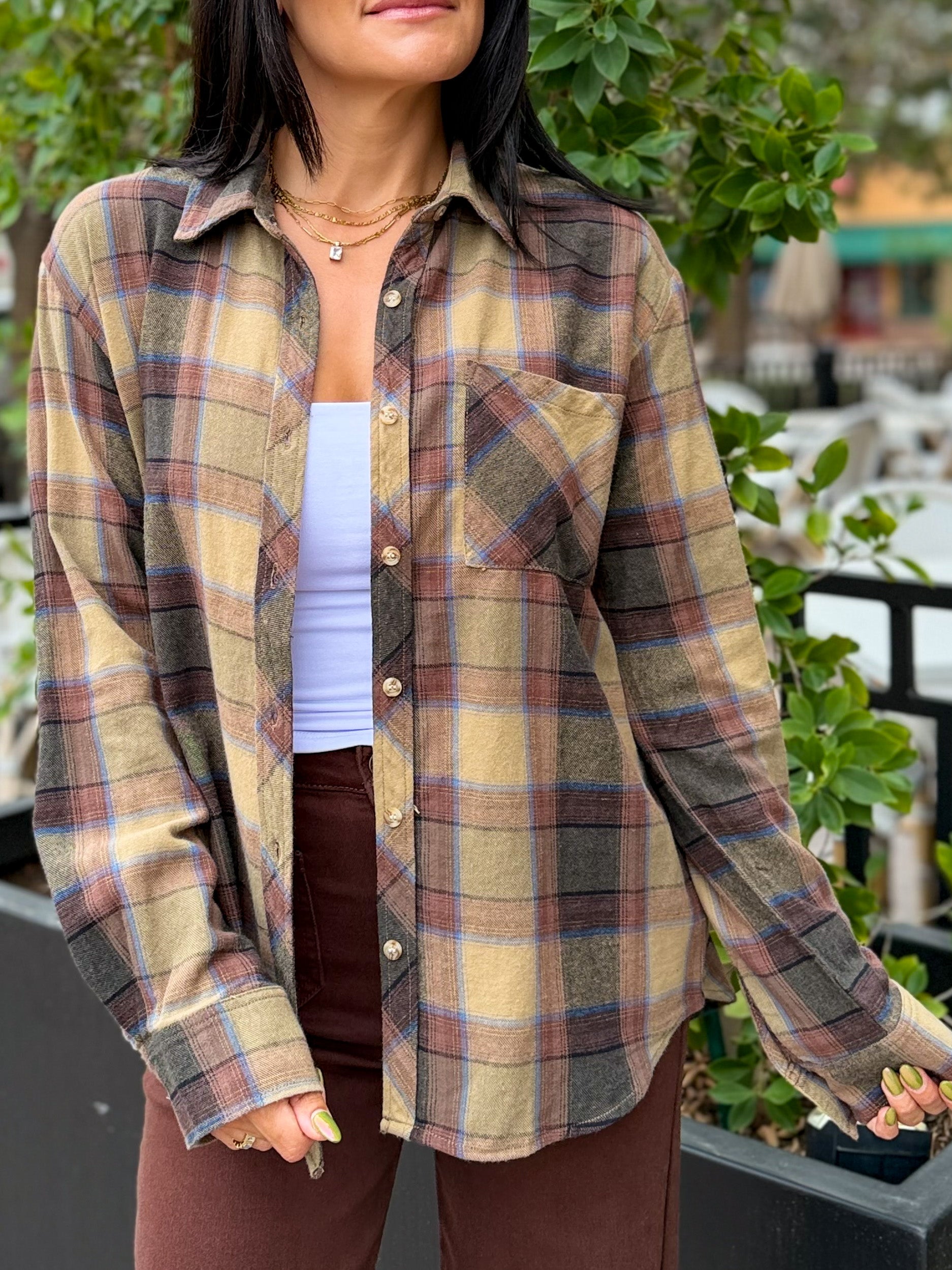 A woman wearing a long-sleeve button-up plaid flannel shirt in beige, brown, and muted blue, layered over a white tank top and brown pants, photographed outdoors with green foliage in the background.