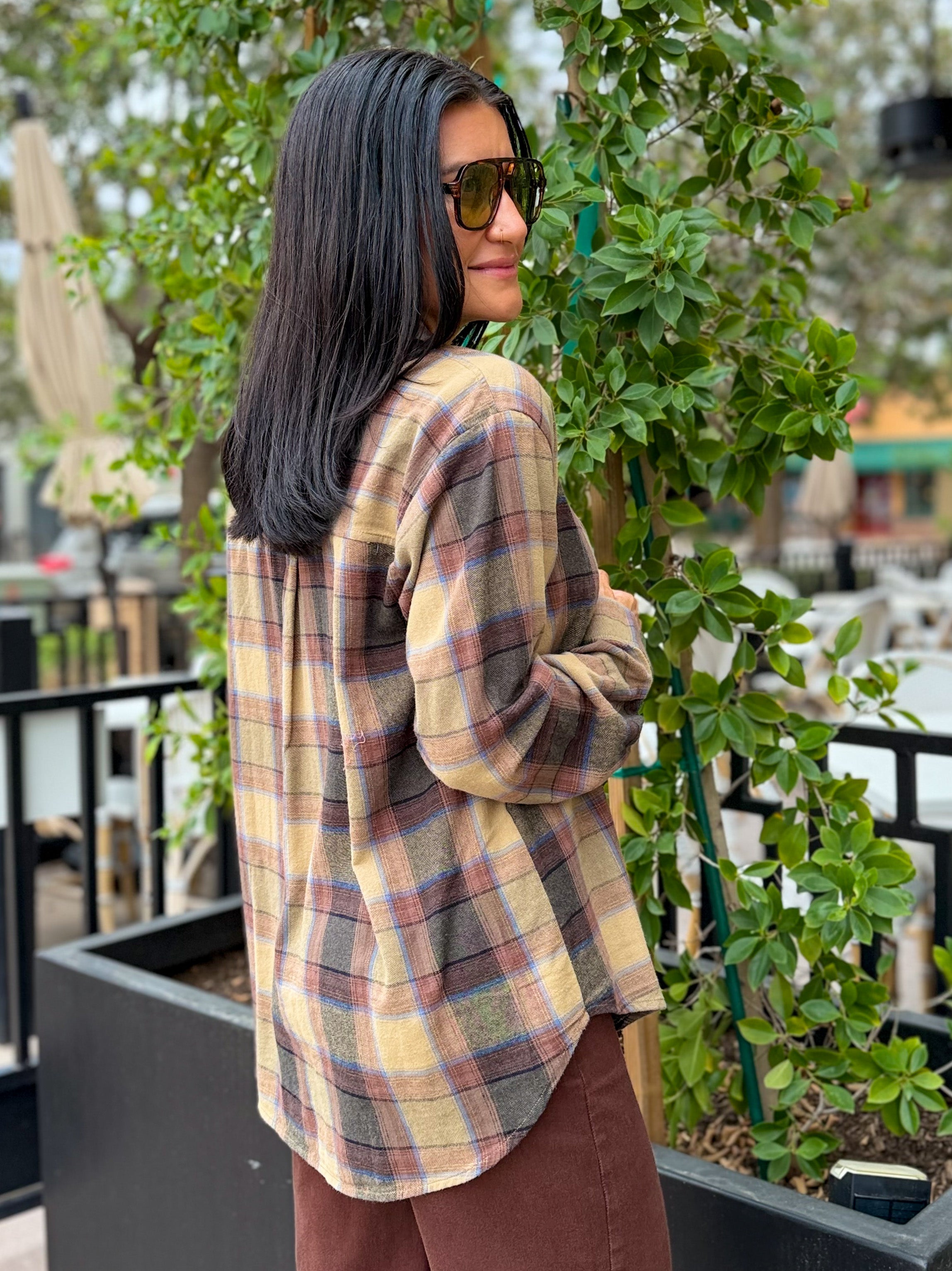 A woman with long black hair and sunglasses, wearing a beige and brown plaid flannel shirt, standing outdoors next to a green plant.