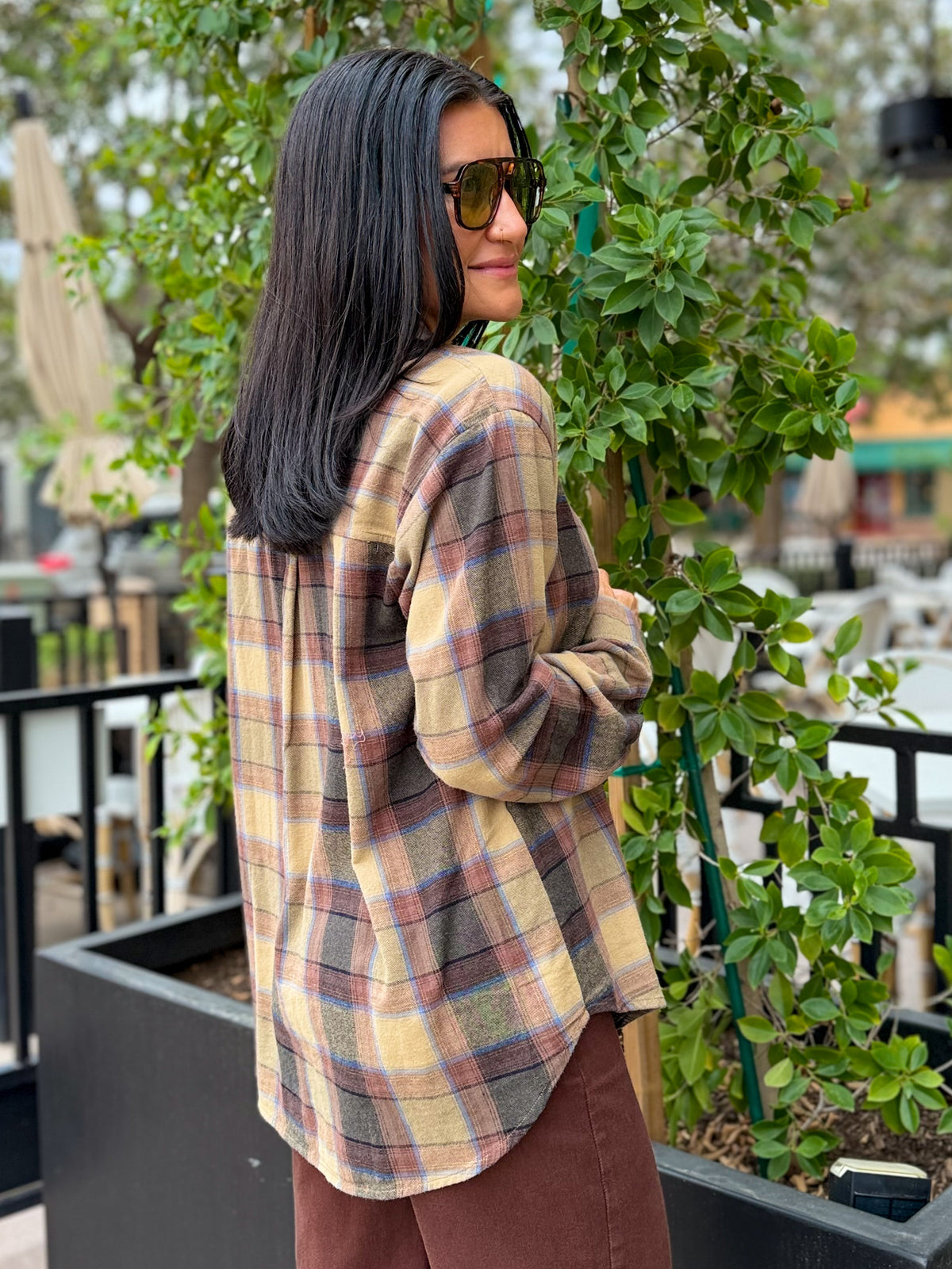 A woman with long black hair and sunglasses, wearing a beige and brown plaid flannel shirt, standing outdoors next to a green plant.