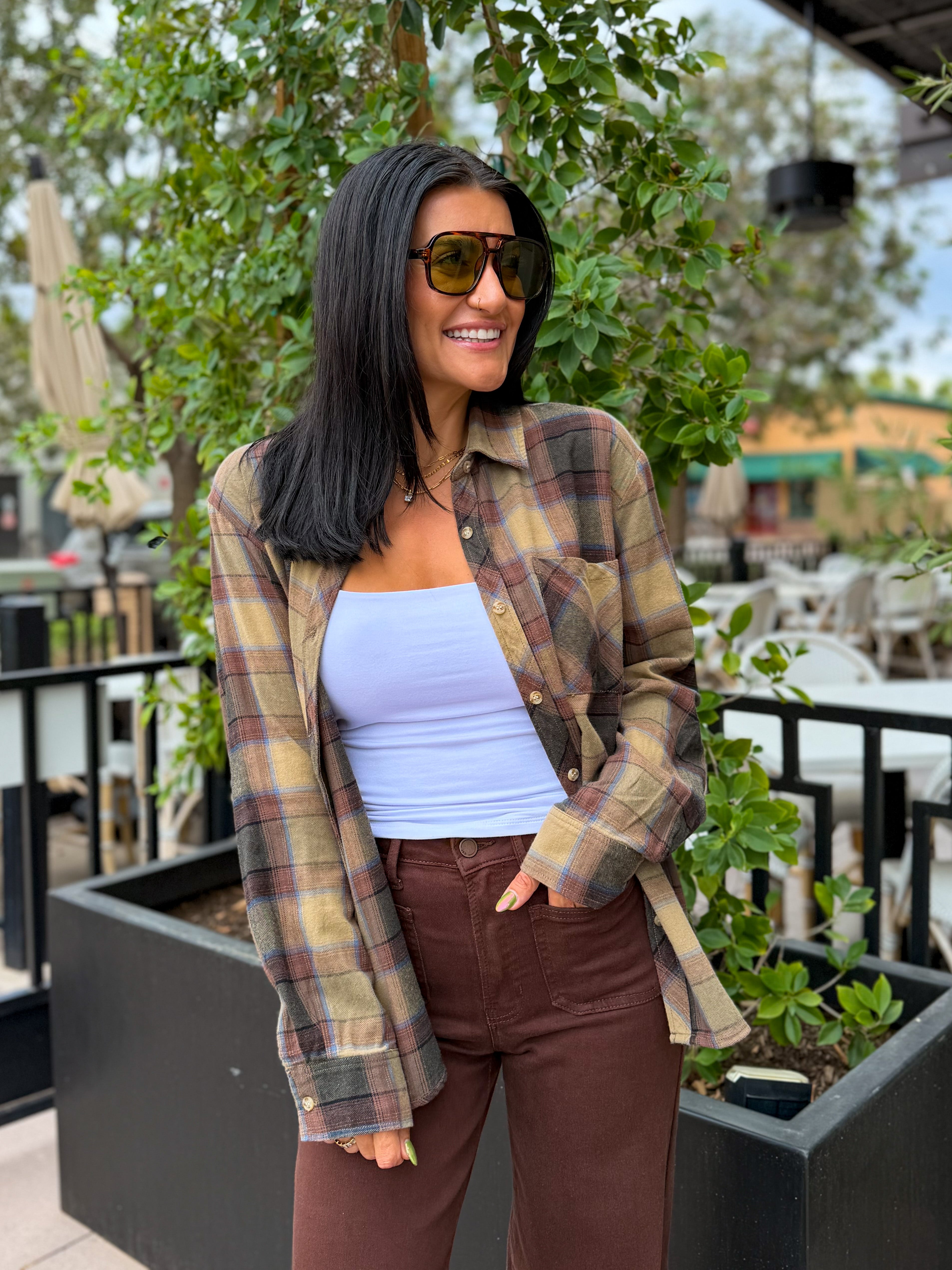 A woman wearing a brown and beige plaid flannel shirt over a light blue tank top and brown pants, standing outdoors in front of green plants.
