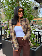 A woman wearing a brown and beige plaid flannel shirt over a light blue tank top and brown pants, standing outdoors in front of green plants.