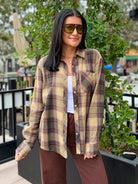 A woman wearing an oversized long-sleeve plaid button-up shirt in beige, brown, and muted red tones, styled over a white top and brown pants, standing outdoors in front of greenery.