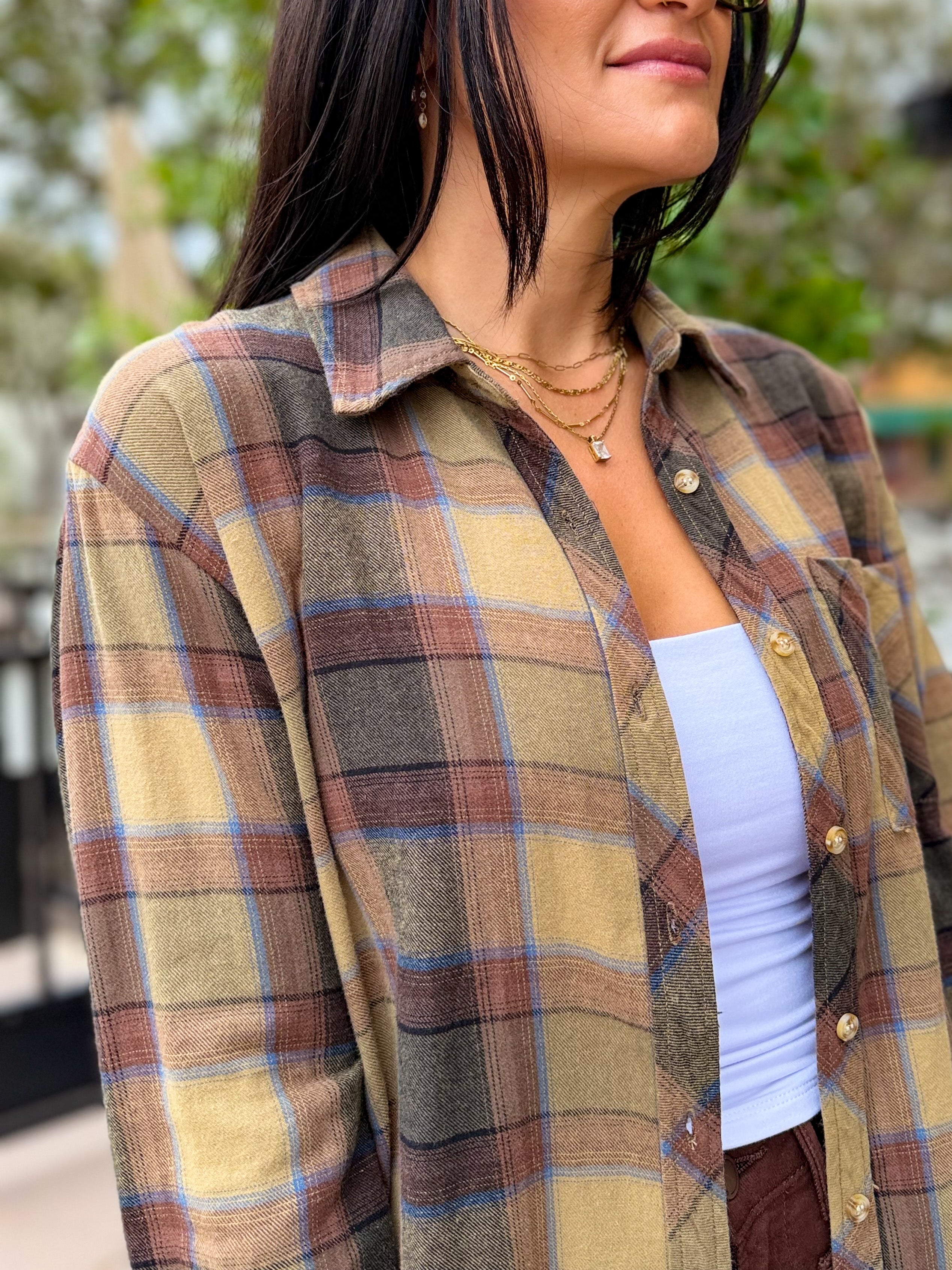 A woman wearing a long-sleeve plaid button-up shirt in muted earthy tones of beige, brown, gray, and light blue, layered over a white tank top.