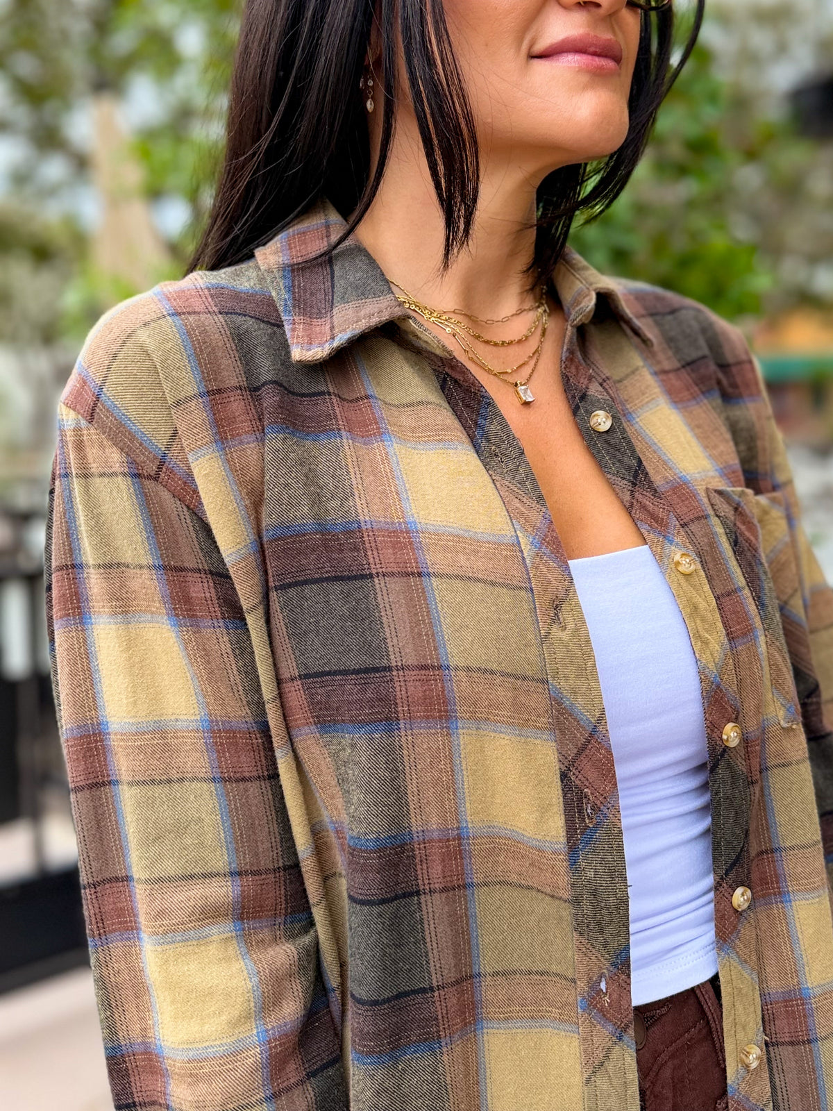 A woman wearing a long-sleeve plaid button-up shirt in muted earthy tones of beige, brown, gray, and light blue, layered over a white tank top.